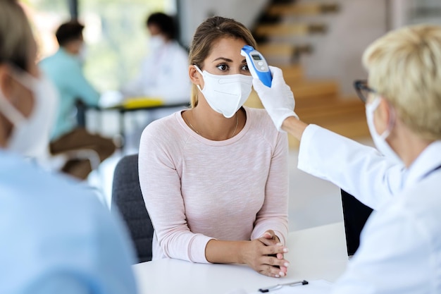 Jeune femme avec masque facial faisant mesurer sa température lors d'un rendez-vous médical à la clinique