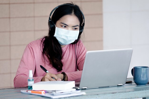 Une jeune femme avec un masque facial travaillant sur un ordinateur portable à la maison pendant la pandémie de coronavirus ou de COVID-19.. Un apprenant ault étudiant en ligne à la maison. Stock photo