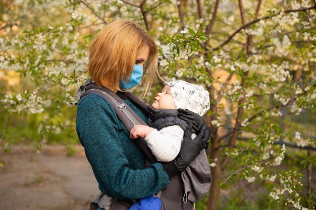 Jeune femme en masque médical avec un enfant dans ses bras arbre fleuri à la fin du printemps