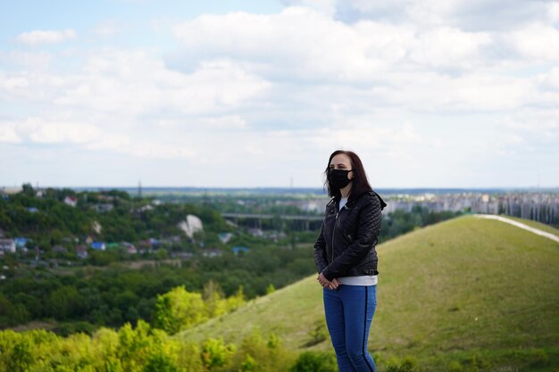 Jeune femme en masque médical noir et veste en cuir sur la colline par temps nuageux Femme adulte marchant seule dans la campagne