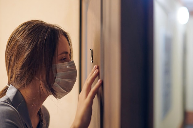 Jeune femme en masque médical à la recherche à travers le judas de la porte d'entrée dans l'appartement lorsque quelqu'un sonne à la porte. Restez à la maison et concept d'auto-isolement