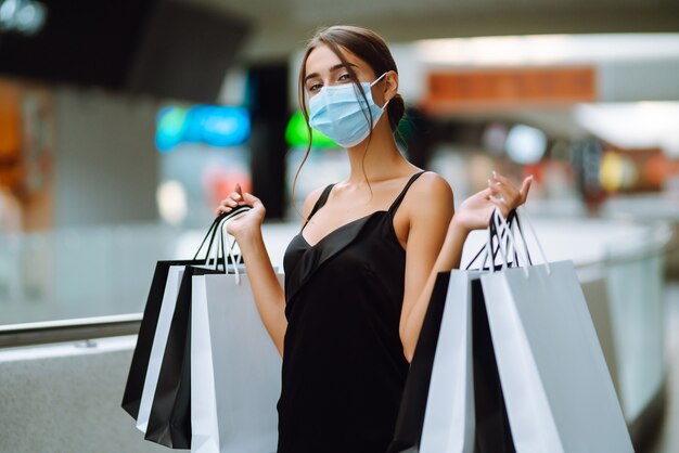 Jeune Femme En Masque Médical Stérile Protecteur Sur Son Visage Avec Des Sacs à Provisions Dans Le Centre Commercial.