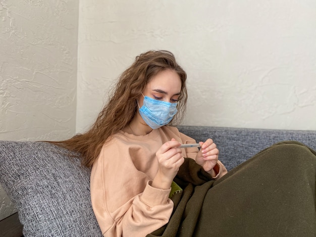 Jeune femme en masque médical avec un thermomètre à la main est assise à la maison sur le canapé