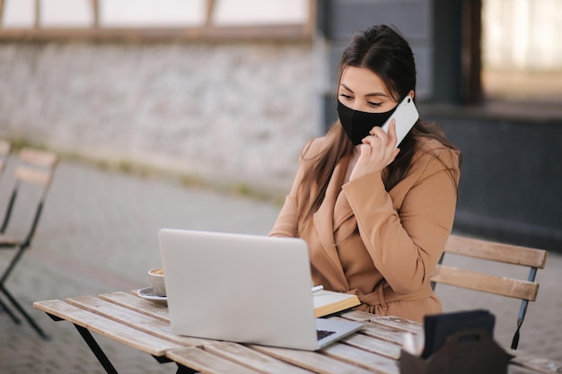 Jeune femme en masque noir assise sur la terrasse d'un café pendant la quarantaine Femme d'affaires travaillant à l'extérieur en quarantaine Femme parlant au téléphone Covid19