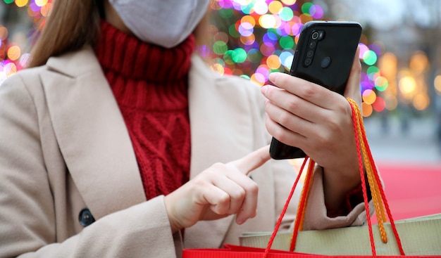 Jeune femme avec masque de protection à l'aide de smartphone pour faire du shopping en ligne et porter des sacs à Noël
