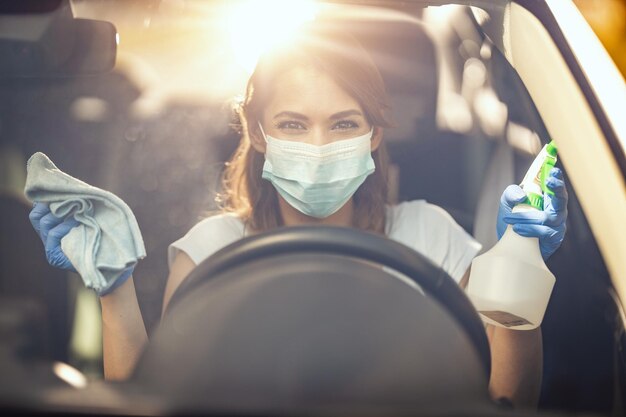 Une jeune femme avec un masque sur le visage et des gants de protection sur ses mains est prête à essuyer sa voiture en tenant un chiffon dans l'une et une bouteille de désinfectant dans l'autre main.