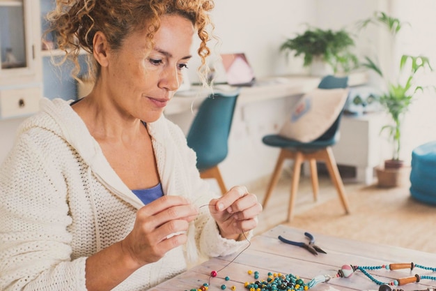 Une jeune femme mature jouissant à la maison avec une activité de loisirs en intérieur faisant des bijoux et des colliers avec des perles colorées et regardant un appareil tablette pour suivre les instructions du tutoriel en ligne