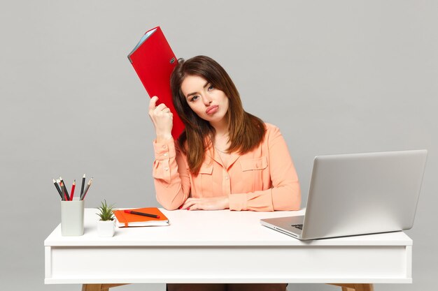 Une jeune femme mécontente dans des vêtements pastel tient un dossier rouge pour les documents de travail au bureau avec un ordinateur portable pc isolé sur fond gris. Concept de style de vie de carrière d'affaires de réalisation. Maquette de l'espace de copie.