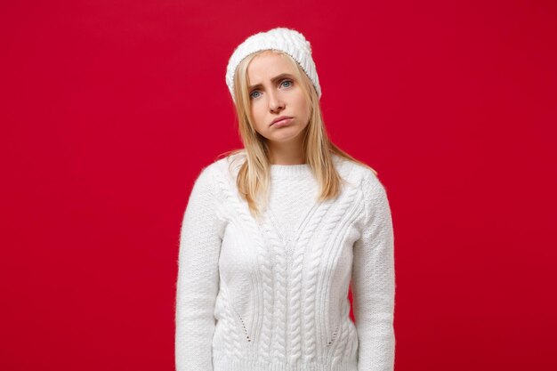 Jeune femme mécontente en pull blanc chapeau isolé sur fond de mur rouge portrait en studio. Mode de vie sain personnes émotions sincères concept de saison froide. Maquette de l'espace de copie. Regarder la caméra.