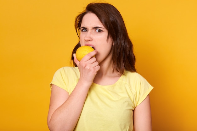Jeune femme mécontente en t-shirt décontracté mordant le citron isolé sur jaune