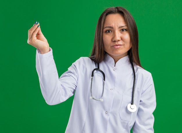 Jeune femme médecin asiatique sans idée portant une robe médicale et un stéthoscope regardant à l'avant faisant un geste d'argent isolé sur un mur vert