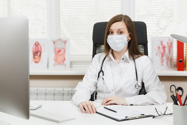 Jeune femme médecin assise au bureau, travaillant sur un ordinateur moderne avec des documents médicaux dans un bureau léger à l'hôpital