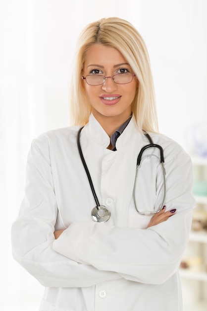 Jeune femme médecin blonde mignonne avec les lunettes, vêtue d'un manteau blanc debout les bras croisés avec un stéthoscope sur le cou, avec un sourire en regardant la caméra.