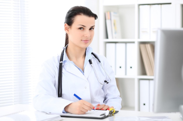 Jeune femme médecin brune assise avec un presse-papiers près de la fenêtre à l'hôpital et remplissant le formulaire d'antécédents médicaux.