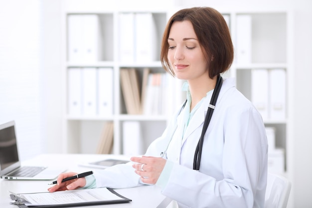 Jeune femme médecin brune assise à la table et travaillant au bureau de l'hôpital. Concept de soins de santé, d'assurance et d'aide. Médecin prêt à examiner le patient