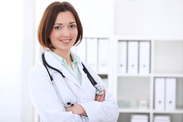 Jeune femme médecin brune debout avec les bras croisés et souriant à l'hôpital