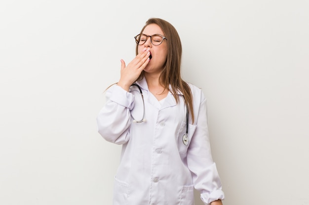 Jeune femme médecin contre un mur blanc bâillant montrant un geste fatigué couvrant la bouche avec la main.