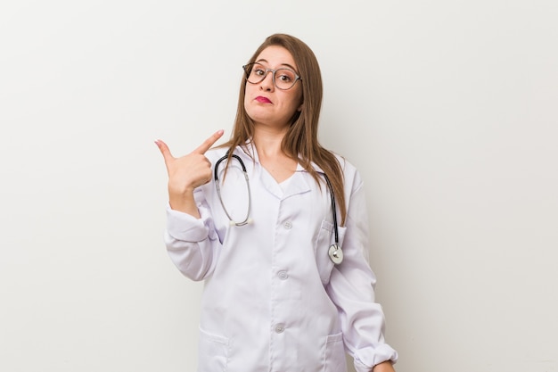 Jeune femme médecin contre un mur blanc pointant du doigt sur vous comme si vous invitiez à vous rapprocher.