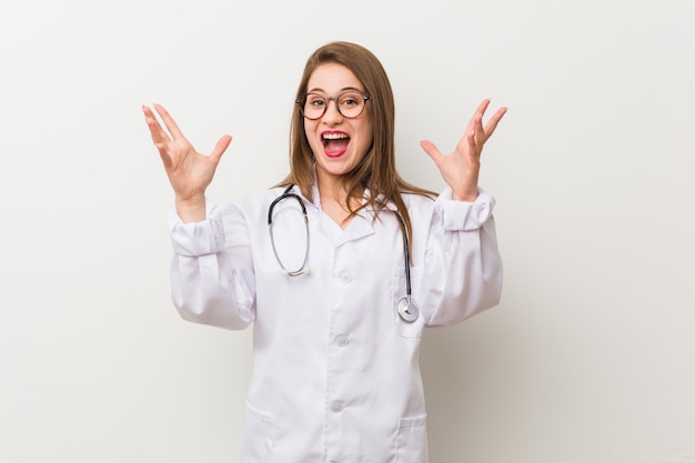 Jeune femme médecin contre un mur blanc recevant une agréable surprise, excitée et levant les mains.