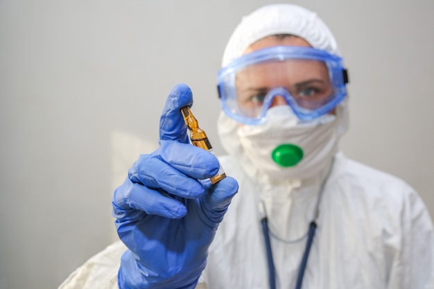 Une jeune femme médecin dans une combinaison de protection tient une ampoule de médicament dans sa main