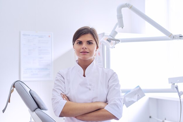 Jeune femme médecin dentiste positive avec les bras croisés, arrière-plan du cabinet de dentisterie