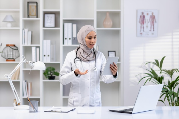 Jeune femme médecin expérimentée en hijab utilisant une tablette pour un appel vidéo, une employée de clinique