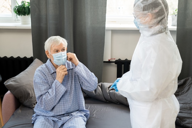 Jeune femme médecin en gants et combinaisons debout devant un homme âgé malade assis sur son lit et mettant un masque de protection sur son visage