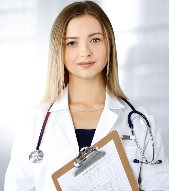 Jeune femme médecin optimiste tient un presse-papiers dans ses mains, tout en se tenant dans une clinique ensoleillée. Portrait d'une femme médecin sympathique avec un stéthoscope. Service médical parfait dans un hôpital. Moi