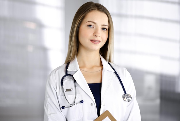 Jeune femme médecin optimiste tient un presse-papiers dans ses mains, tout en se tenant dans une clinique. Portrait d'une femme médecin sympathique avec un stéthoscope. Service médical parfait dans un hôpital. Médecine