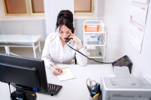 Jeune femme médecin, parler par téléphone et écrire quelque chose dans son bureau