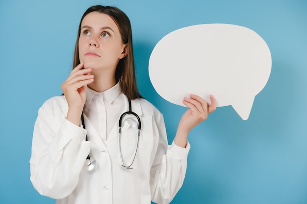 Photo jeune femme médecin pensive en blouse blanche et stéthoscope tenir vide vide dites bulle de dialogue nuage, isolée sur fond bleu. concept de virus de la grippe pandémique coronavirus 2019-ncov sars covid-19