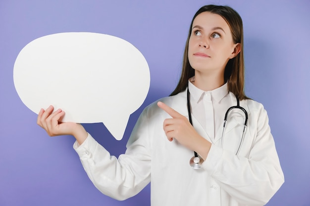 Photo jeune femme médecin pensive en stéthoscope de robe médicale tenir vide vide dites bulle de dialogue nuage, posant isolé sur mur de fond violet studio. concept de médecine de santé du personnel de santé