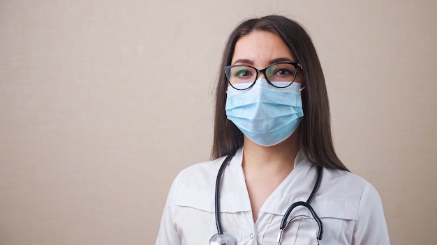 Jeune femme médecin qualifiée avec des lunettes et un masque jetable bleu en robe blanche pose pour la caméra debout sur fond beige vue rapprochée