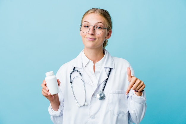 Jeune femme médecin russe tenant la bouteille de pilules sur les points bleus vers le bas avec les doigts, sentiment positif