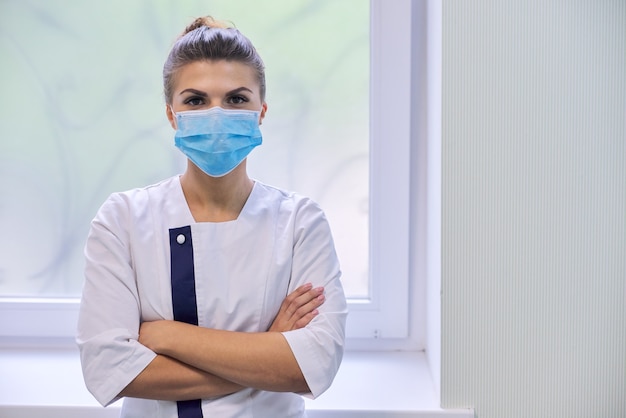 Jeune femme médecin sérieuse confiante en masque médical avec les bras croisés regardant la caméra, le mur de l'hôpital