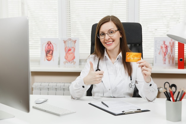 Jeune femme médecin souriante assise au bureau, tenant une carte de crédit, travaillant avec des documents médicaux dans un bureau léger à l'hôpital