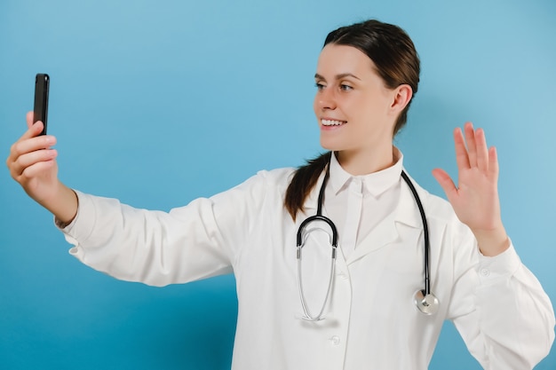 Une jeune femme médecin souriante tenant une conversation téléphonique avec un patient fait une conférence vidéo en ligne de télémédecine, isolée sur un mur bleu. Application de chat de soins de santé mobile virtuel de télémédecine de consultation à distance Gp