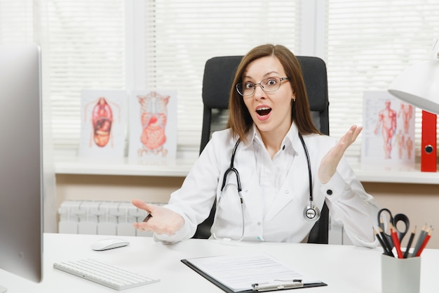 Jeune femme médecin surprise assise au bureau travaillant sur ordinateur avec des documents médicaux dans un bureau léger à l'hôpital