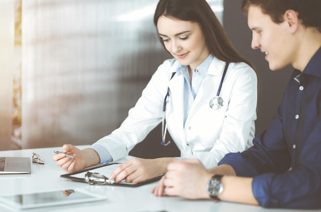 Une jeune femme médecin sympathique écoute sa patiente, assise ensemble dans une clinique ensoleillée. Concepts de médecine et de soins de santé.