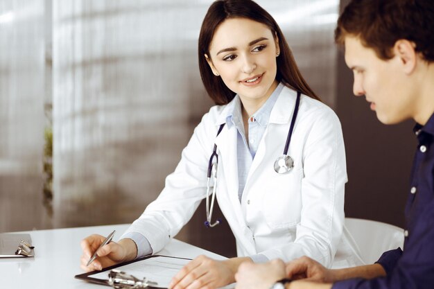 Photo une jeune femme médecin sympathique écoute son patient alors qu'ils sont assis ensemble au bureau dans un cabinet. le médecin tient un presse-papiers dans ses mains