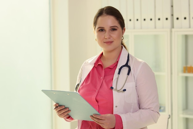 Jeune femme médecin sympathique tenant un presse-papiers avec un stylo de documents médicaux et regardant la caméra