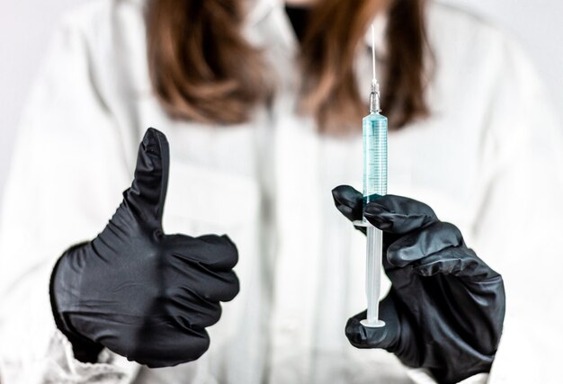 Jeune femme médecin tenant la seringue et soulever le pouce avec des gants noirs avec robe, concept de succès de la médecine et du vaccin.