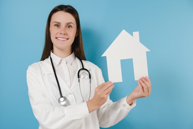 Jeune femme médecin en uniforme blanc médical professionnel et stéthoscope détient une petite maison de papier, regardant la caméra en souriant, posant isolé sur fond bleu. Concept d'assurance-vie de soins de santé