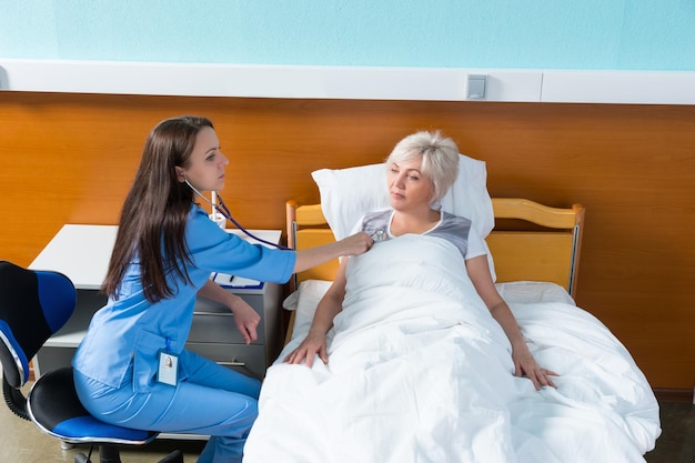 Jeune femme médecin en uniforme avec phonendoscope écoutant le rythme cardiaque d'un patient assis sur une chaise près d'un lit d'hôpital dans la salle d'hôpital. Concept de soins de santé