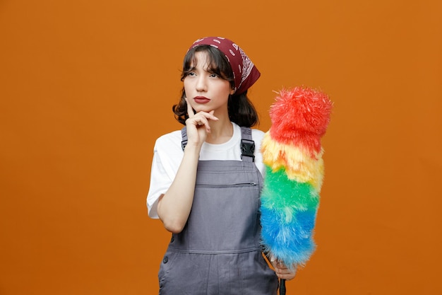 Jeune femme de ménage réfléchie portant l'uniforme et le bandana tenant un plumeau regardant de côté tout en gardant la main sur le menton isolé sur fond orange