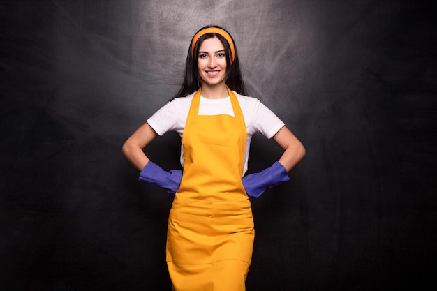 Jeune femme de ménage souriante et confiante en tablier jaune et gants en caoutchouc bleu gardant les mains sur la taille et regardant la caméra sur fond noir