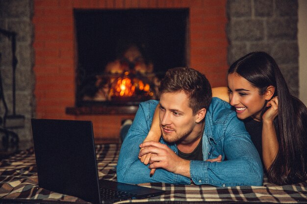 Jeune, femme, mensonge, ensemble, couverture, plancher Ils regardent un film sur un ordinateur portable. Le couple est heureux. Ils sont près de la cheminée.