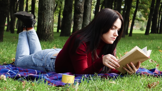 Une jeune femme ment et lit son livre préféré sur une couverture ou une couverture à carreaux en laine dans un parc de la ville sur l'herbe verte par une agréable journée ensoleillée. Le concept de récréation et de loisirs, d'éducation et d'étude.