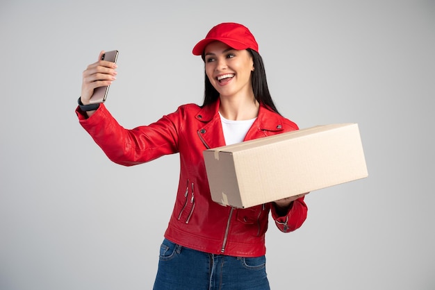 Jeune femme de messagerie brune portant un chapeau rouge et un uniforme faisant du selfie sur le smartphone avec un colis de placard sur un fond isolé avec un visage heureux