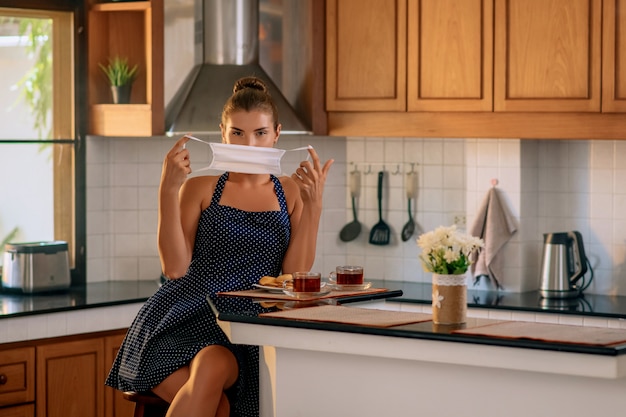 Une jeune femme met un masque médical protecteur sur son visage alors qu'elle était assise dans une cuisine confortable. Auto-isolement. Reste à la maison. Quarantaine